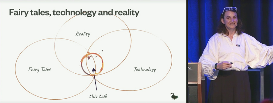 Tea Uglow stands next to a slide showing three intersecting circile labeled Fairy Tales, Reality, and Technology. At their intersection is a circle formed by a coffee cup stain labelled This Talk. 