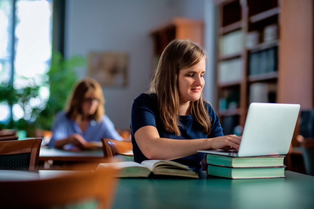 Woman using a laptop in a library or study space, smiling as she works with books stacked beside her.