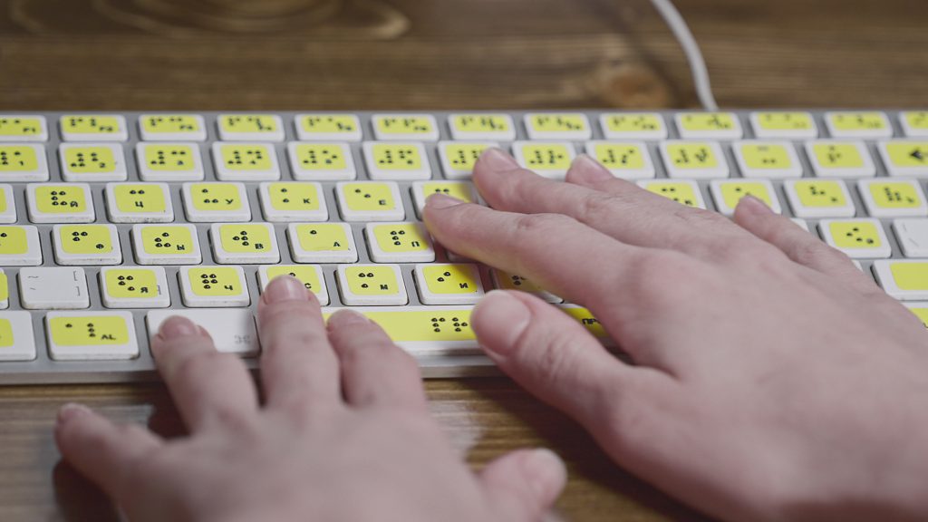 Visually impaired user’s hands moving across computer keyboard with braille labels.