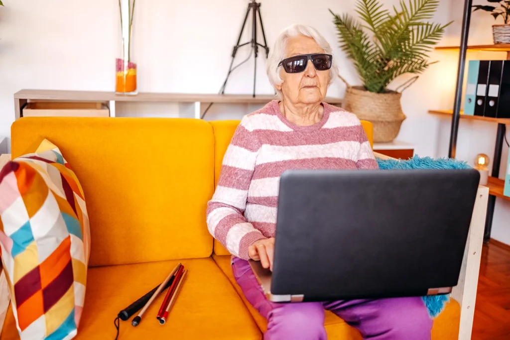 Older blind woman sitting on a sofa with a laptop on her lap.
