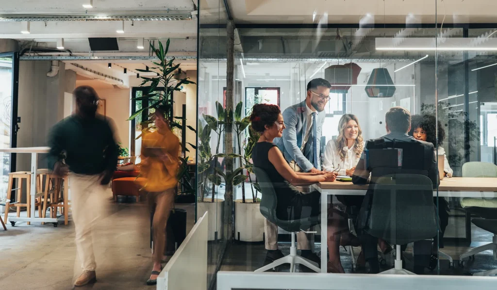 Business meeting with several participants in glass conference room and 2 colleagues outside walking by.
