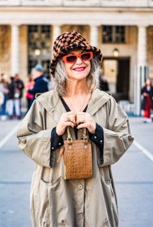 Smiling woman with gray hair standing in a plaza wearing red glasses, checkered hat and holding a small, brown purse.