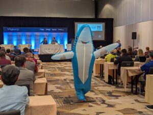 A human in a smiling life-size blue and white shark outfit walks down the center aisle of the room.
